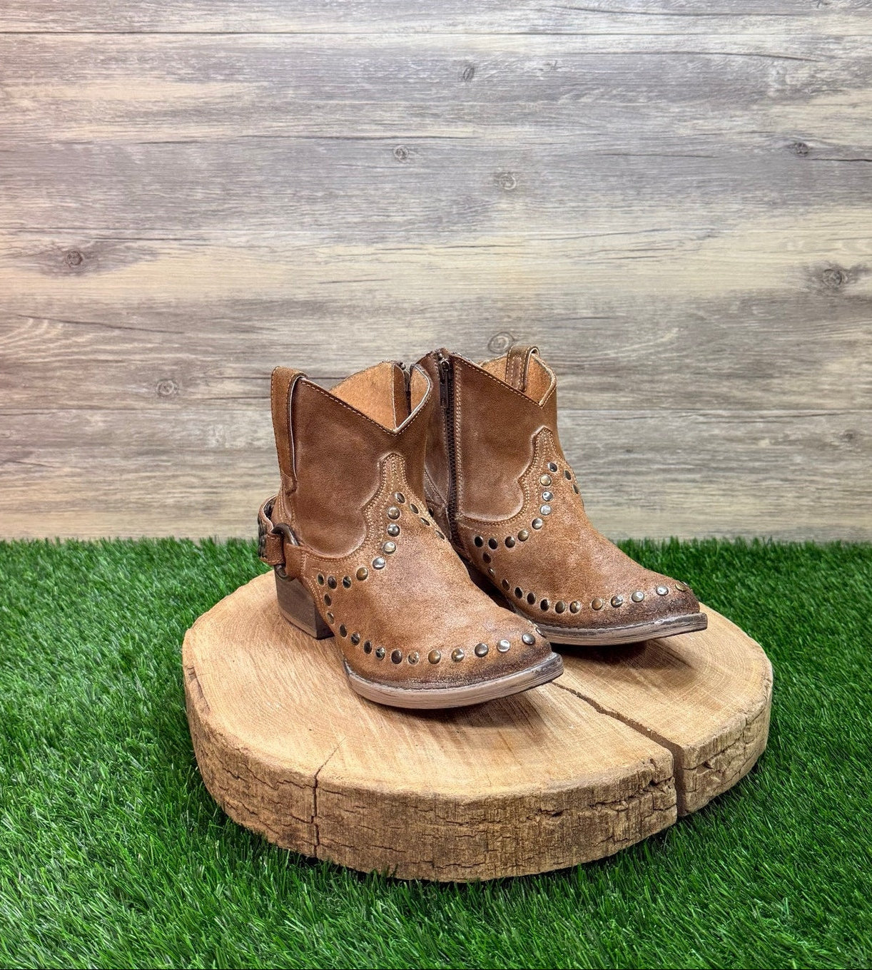 Pair of brown ankle studded cowboy boots with studs on a wooden platform against a wooden wall.