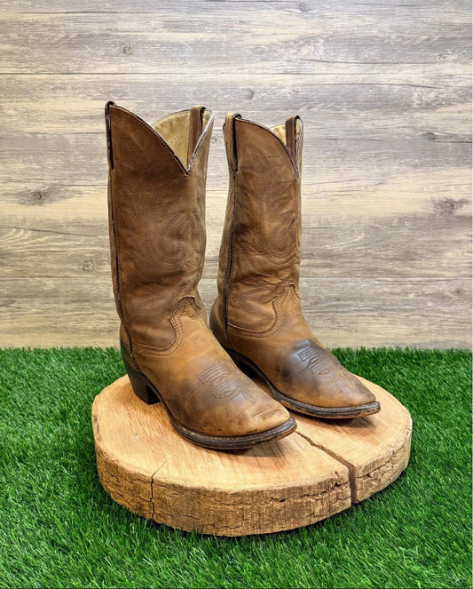 Pair of brown cowboy boots on a wooden block with a grass and wooden panel background
